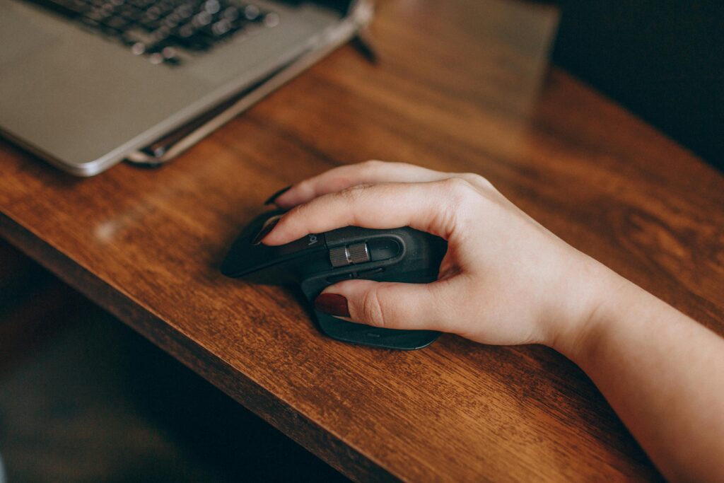 A close-up image of a hand with manicured nails using a wireless mouse on a wooden desk beside a laptop.